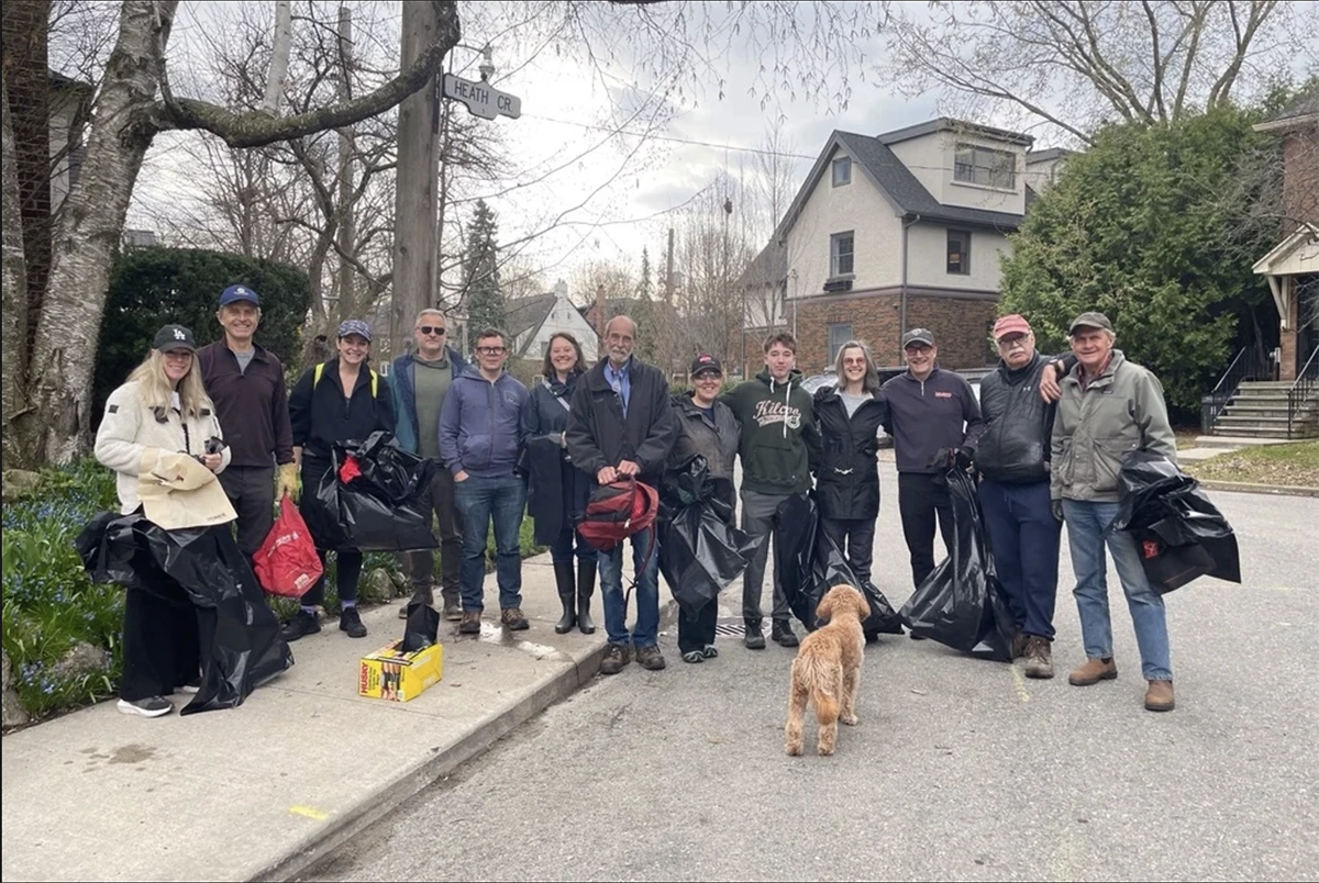 A group of adults on a gray, cold spring day, standing on a sidewalk and street in a residential neighbourhood carrying garbage bags