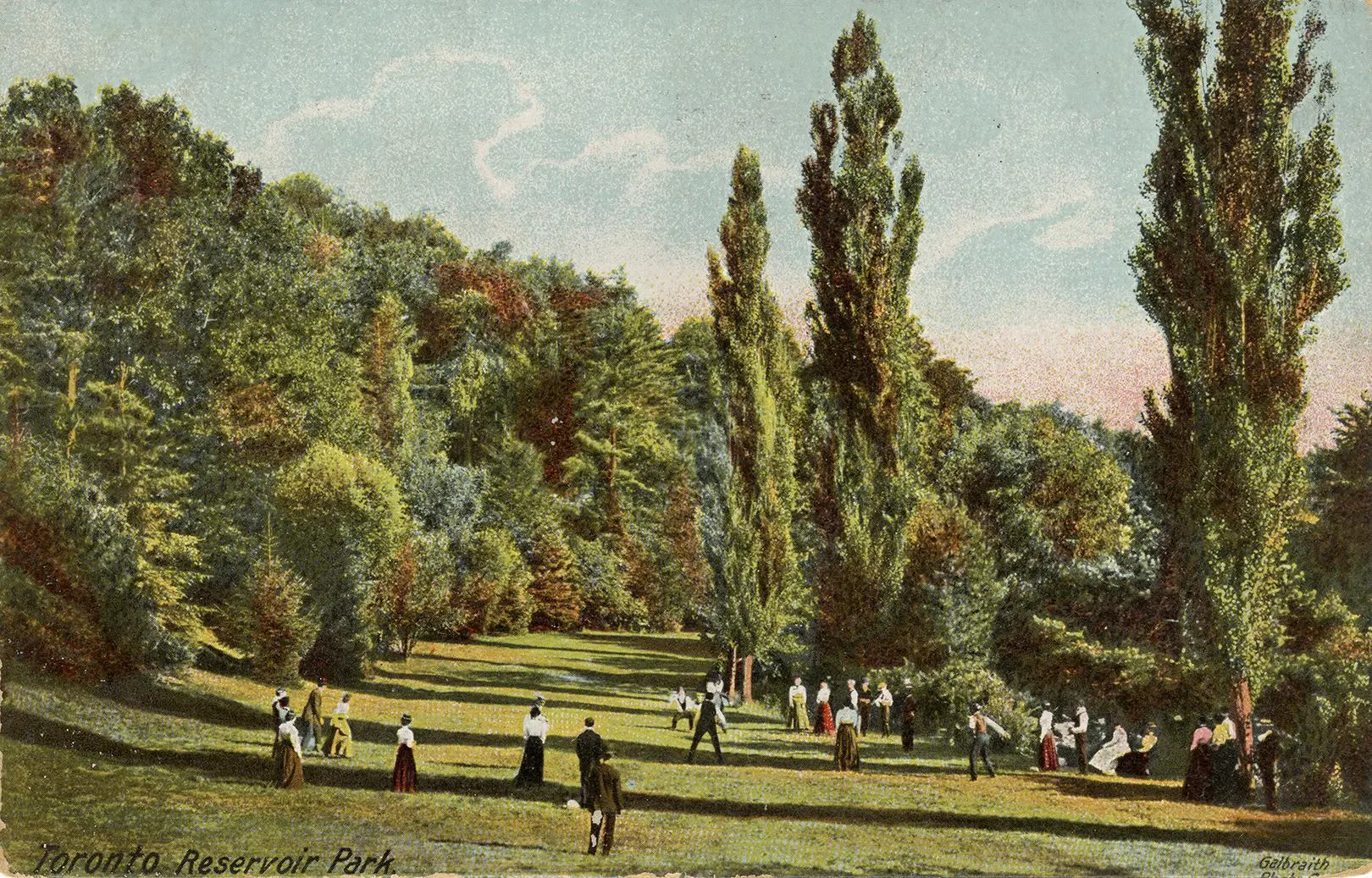 People in early 20th c. clothing scattered over grassy area with treed hillside in background