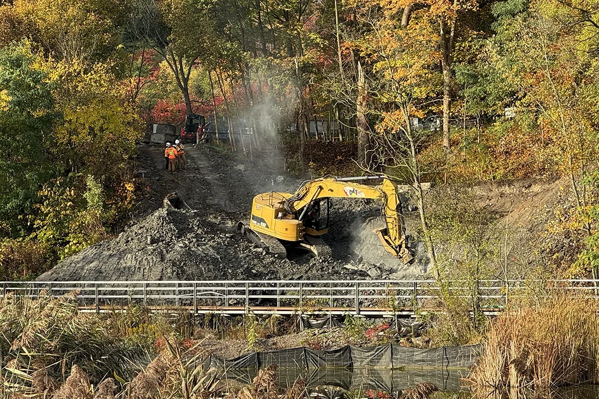 Yellow excavator digging in wooded ravine with pedestrian footbridge in foreground