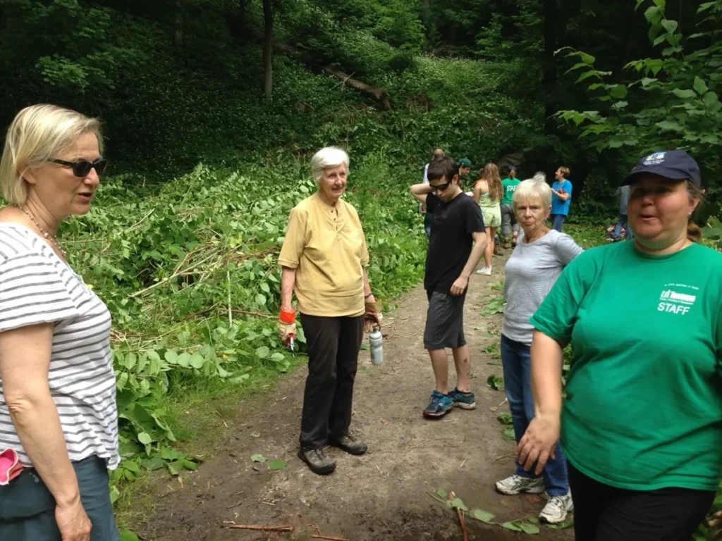 A group of hikers pauses on a wooded trail, with a woman in a green shirt and navy cap in the foreground.