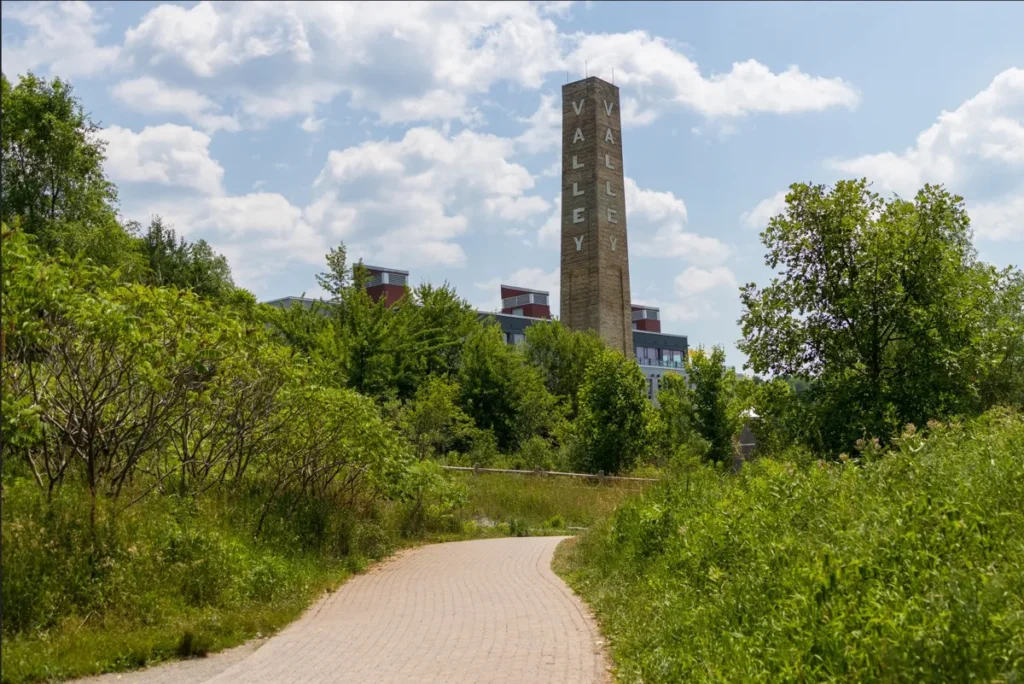 A paved path winds through green vegetation toward a tall stone obelisk beneath a partly cloudy sky.