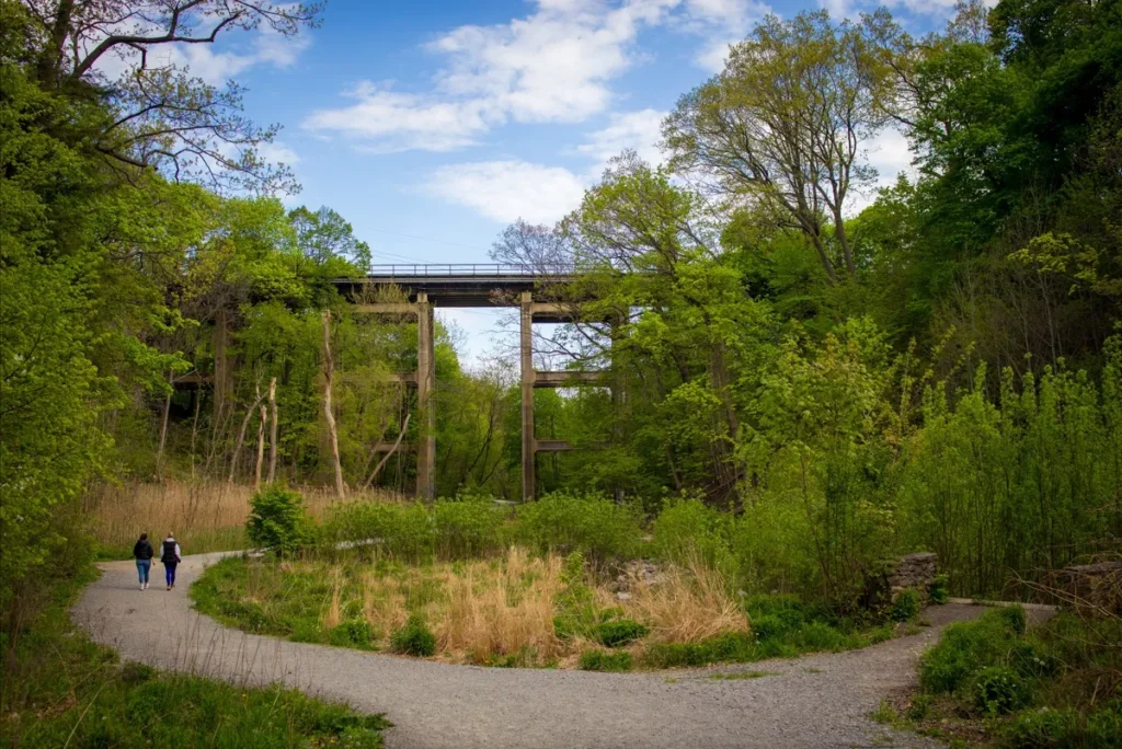 A gravel path winds through a green park toward a tall bridge, with two people walking beneath it.