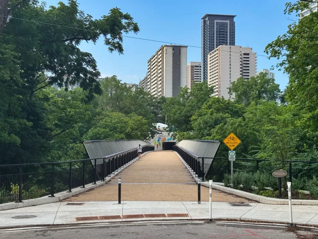 A pedestrian bridge leads through trees toward several high-rise buildings under a clear blue sky.