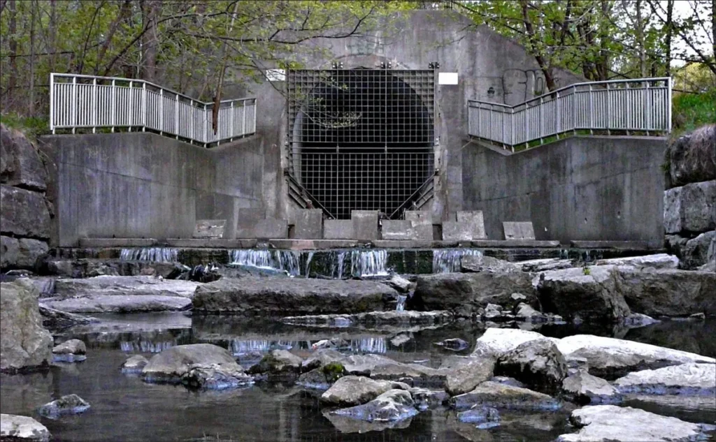 A concrete water outlet with a metal grate releases water over rocky steps into a stream.