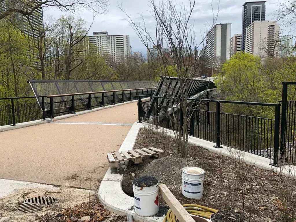 A paved pedestrian path crosses a black metal bridge through a park, with construction buckets and materials in the foreground and city buildings beyond.