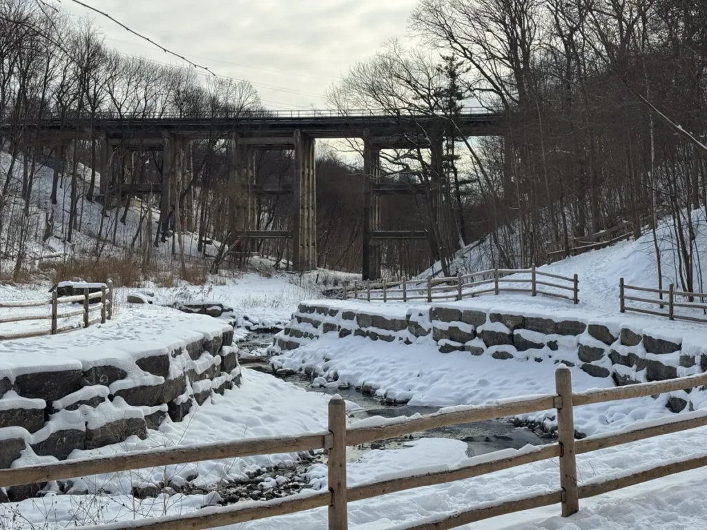A snow-covered stone structure and wooden fences sit beneath a tall bridge in a leafless winter forest.