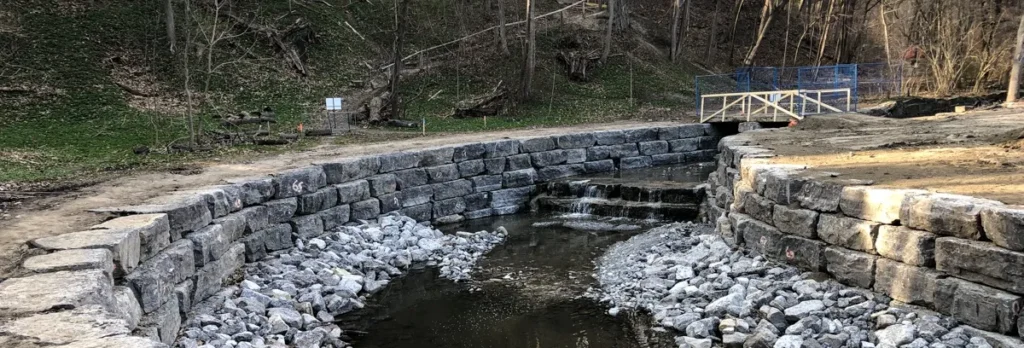 A stone-lined creek runs beneath a small blue footbridge, with terraced stone walls along both banks.