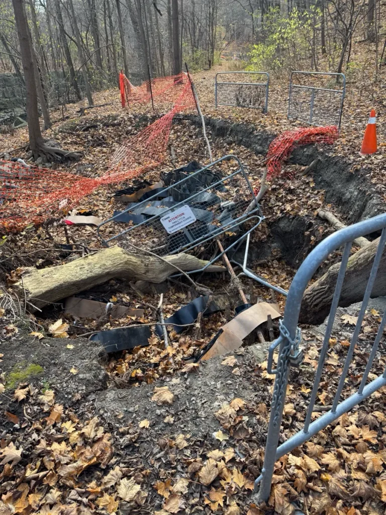 A fenced-off construction excavation with exposed pipes, debris, and a metal barrier in a wooded area.