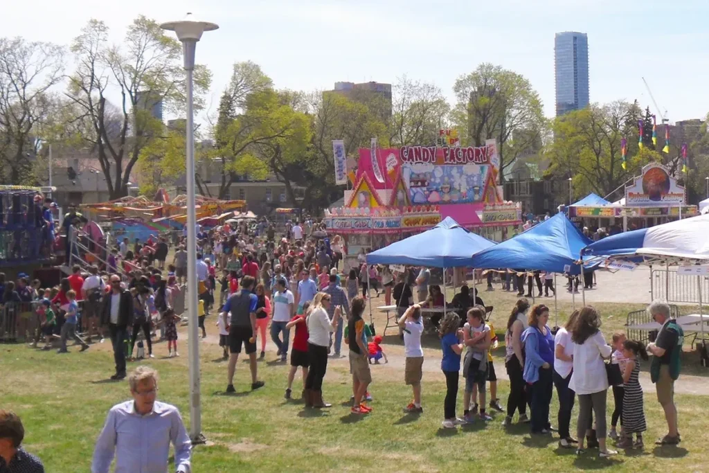 Crowds gather at an outdoor festival with colorful tents and a tall blue skyscraper in the background.