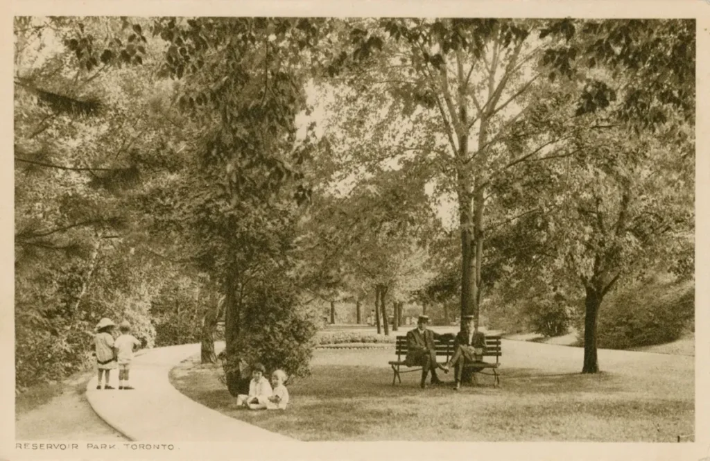 Sepia photograph of a horse-drawn carriage beneath trees in a park, with people walking along a path.