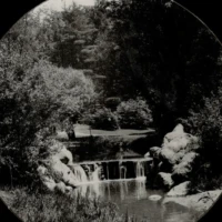 A black-and-white circular photograph of a small waterfall flowing through a wooded landscape.