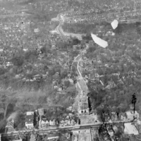Black-and-white aerial photograph of a city with a long road running through it and several plumes of smoke rising in the distance.