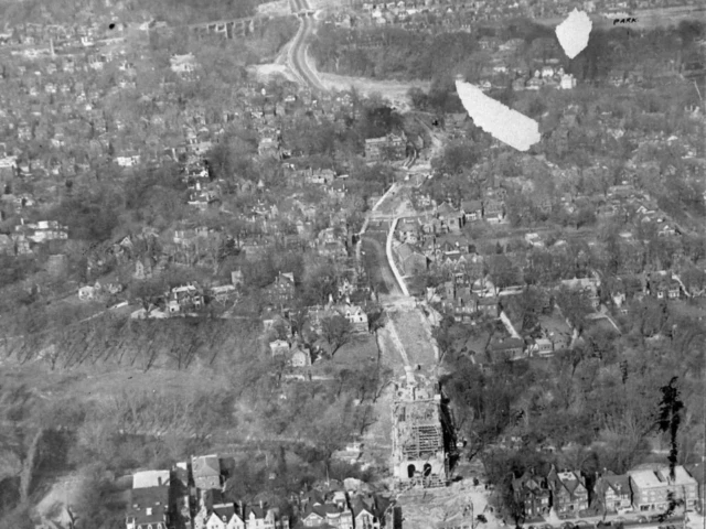 Black-and-white aerial photograph of a city with a long road running through it and several plumes of smoke rising in the distance.