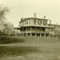 Sepia photograph of a large two-story house with a wraparound porch on a grassy hill, surrounded by trees.