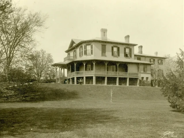 Sepia photograph of a large two-story house with a wraparound porch on a grassy hill, surrounded by trees.