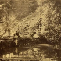 Sepia photograph of people gathered beside a pond beneath a rocky, wooded hillside.