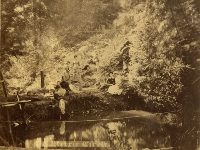 Sepia photograph of people gathered beside a pond beneath a rocky, wooded hillside.