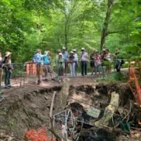 A group of people watches from behind a railing as workers inspect a large sinkhole in a wooded area.