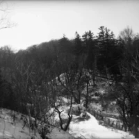 A black-and-white winter landscape with a winding snowy path through a wooded valley.