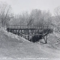 A wooden bridge crosses a snowy ravine among bare trees.