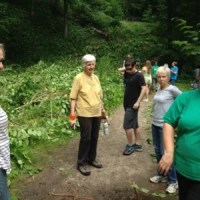 A group of hikers pauses on a wooded trail, with a woman in a green shirt and navy cap in the foreground.