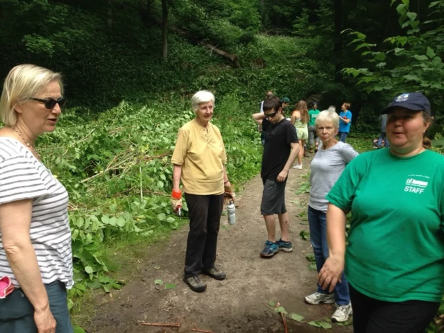 A group of hikers pauses on a wooded trail, with a woman in a green shirt and navy cap in the foreground.