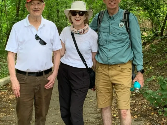 Three hikers stand together on a wooded trail, wearing outdoor clothes and hats.