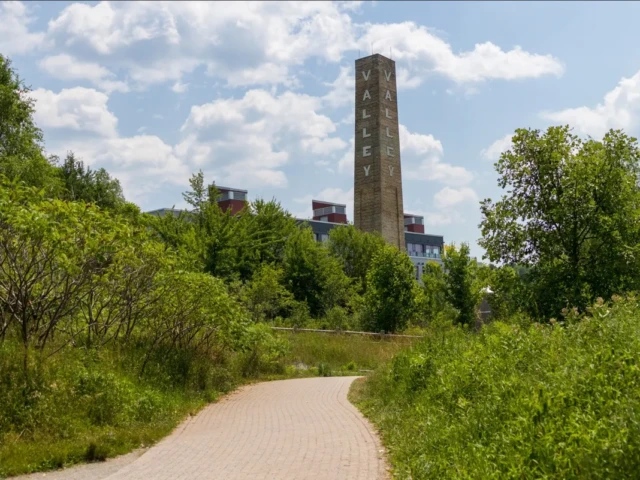 A paved path winds through green foliage toward a tall stone obelisk beneath a cloudy sky.
