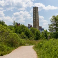 A paved path winds through green vegetation toward a tall stone obelisk beneath a partly cloudy sky.
