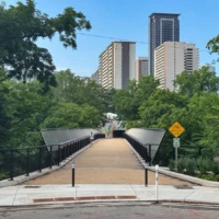 A pedestrian bridge leads through trees toward several high-rise buildings under a clear blue sky.