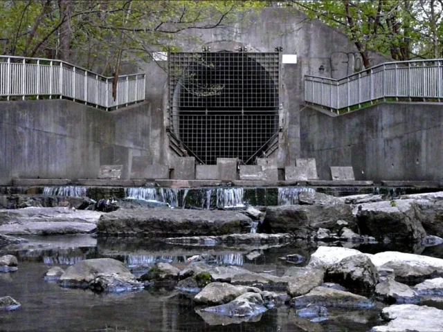 A concrete water outlet with a metal grate releases water over rocky steps into a stream.
