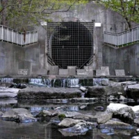 A concrete water outlet with a metal grate releases water over rocky steps into a stream.