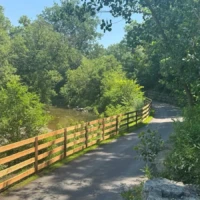 A paved trail curves beside a wooden railing through lush green trees and vegetation.