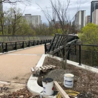 A paved pedestrian path crosses a black metal bridge through a park, with construction buckets and materials in the foreground and city buildings beyond.