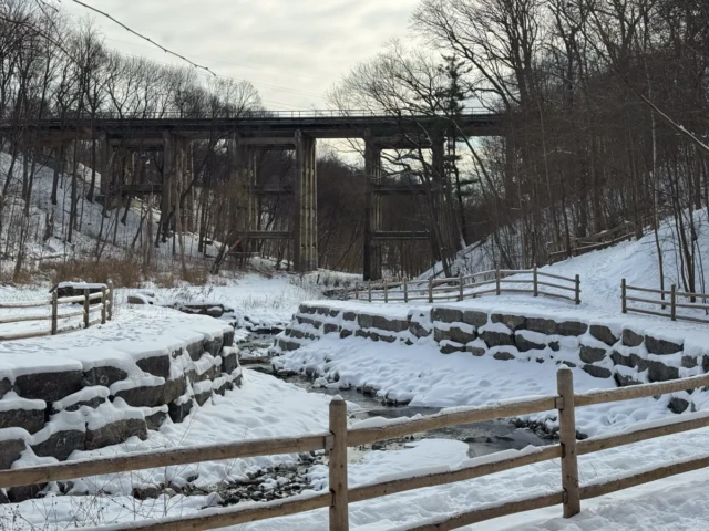 A snow-covered stone terrace and wooden fences sit beneath a tall, weathered trestle bridge in a leafless forest.