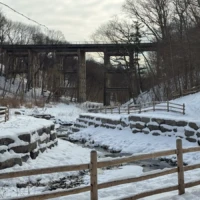 A snow-covered stone terrace and wooden fences sit beneath a tall, weathered trestle bridge in a leafless forest.