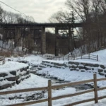 A snow-covered stone terrace and wooden fences sit beneath a tall, weathered trestle bridge in a leafless forest.