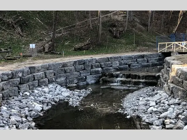 A stone-lined creek runs beneath a small blue footbridge, with terraced stone walls along both banks.