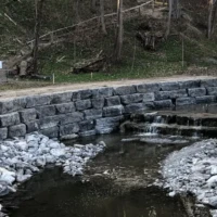 A stone-lined creek runs beneath a small blue footbridge, with terraced stone walls along both banks.