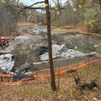 A wooded construction site with an excavated, water-filled area enclosed by orange safety fencing and construction equipment.