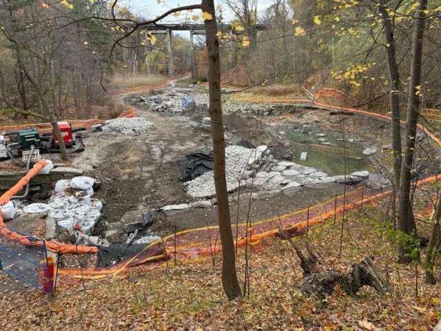 A wooded construction site with a muddy excavation pit, stone fill, orange safety fencing, and construction equipment.