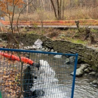 A small stream flows through a stone-lined channel behind a blue metal fence, with orange safety barriers and autumn trees nearby.