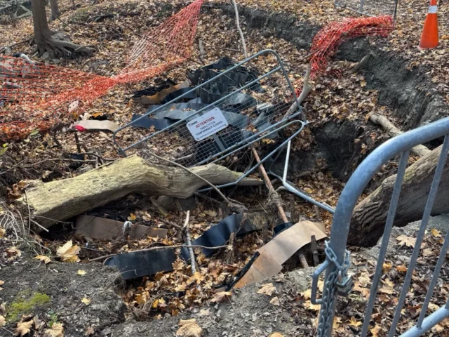 A fenced-off construction excavation with exposed pipes, debris, and a metal barrier in a wooded area.