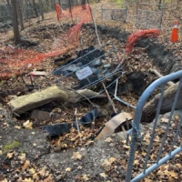 A fenced-off construction excavation with exposed pipes, debris, and a metal barrier in a wooded area.