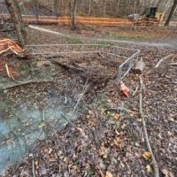 A muddy, leaf-covered construction area is cordoned off with metal barriers and orange safety fencing.