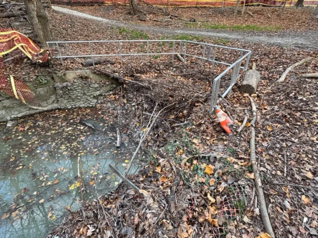 A muddy, leaf-covered construction area is cordoned off with metal barriers and orange safety fencing.