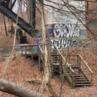 Graffiti-covered concrete structure and wooden stairways among leafless trees in a wooded area.