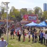 Crowds gather at an outdoor festival with colorful tents and a tall blue skyscraper in the background.