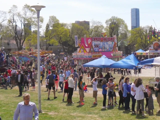 Crowds gather at an outdoor festival with colorful tents and a tall blue skyscraper in the background.