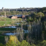 A green park with winding ponds and trees below a hilltop bridge and buildings.